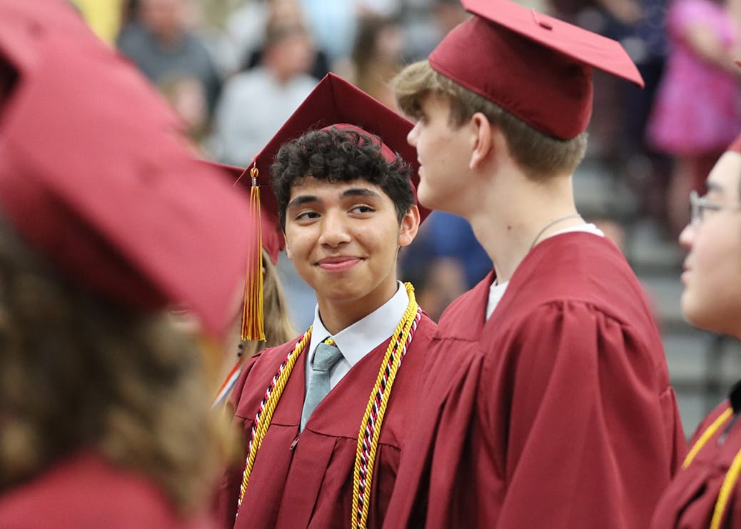 Students in maroon caps and gowns smile and talk during a graduation ceremony, with one student wearing honor cords and looking at a classmate in the crowd-filled gymnasium.